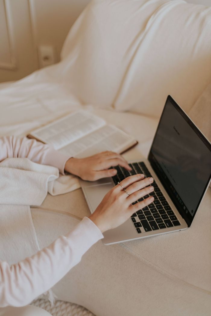 A cozy indoor setting with hands typing on a laptop, next to an open book on a soft surface.
