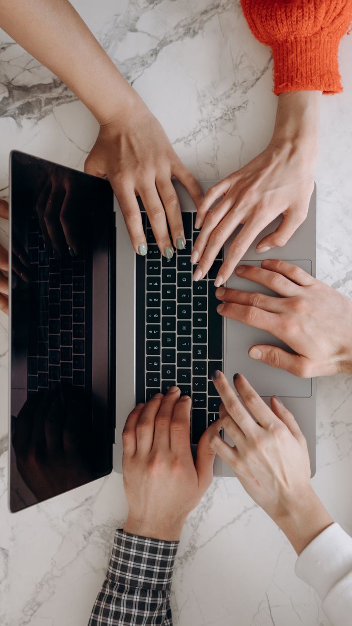 A top-down view of multiple hands typing on a laptop keyboard on a marble desk, indicating teamwork.